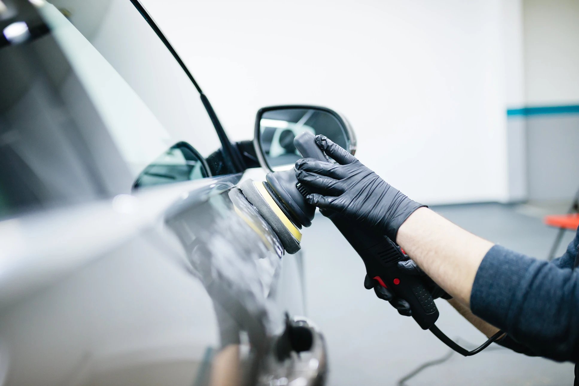 Intérieur d'atelier automobile, technicien en gants noirs utilisant une polisseuse orbitale électrique sur carrosserie blanche de véhicule