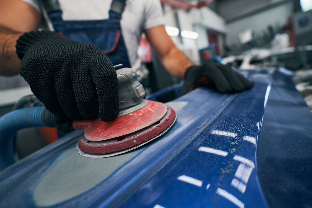 Atelier de carrosserie automobile avec technicien utilisant une ponceuse orbitale sur carrosserie bleue