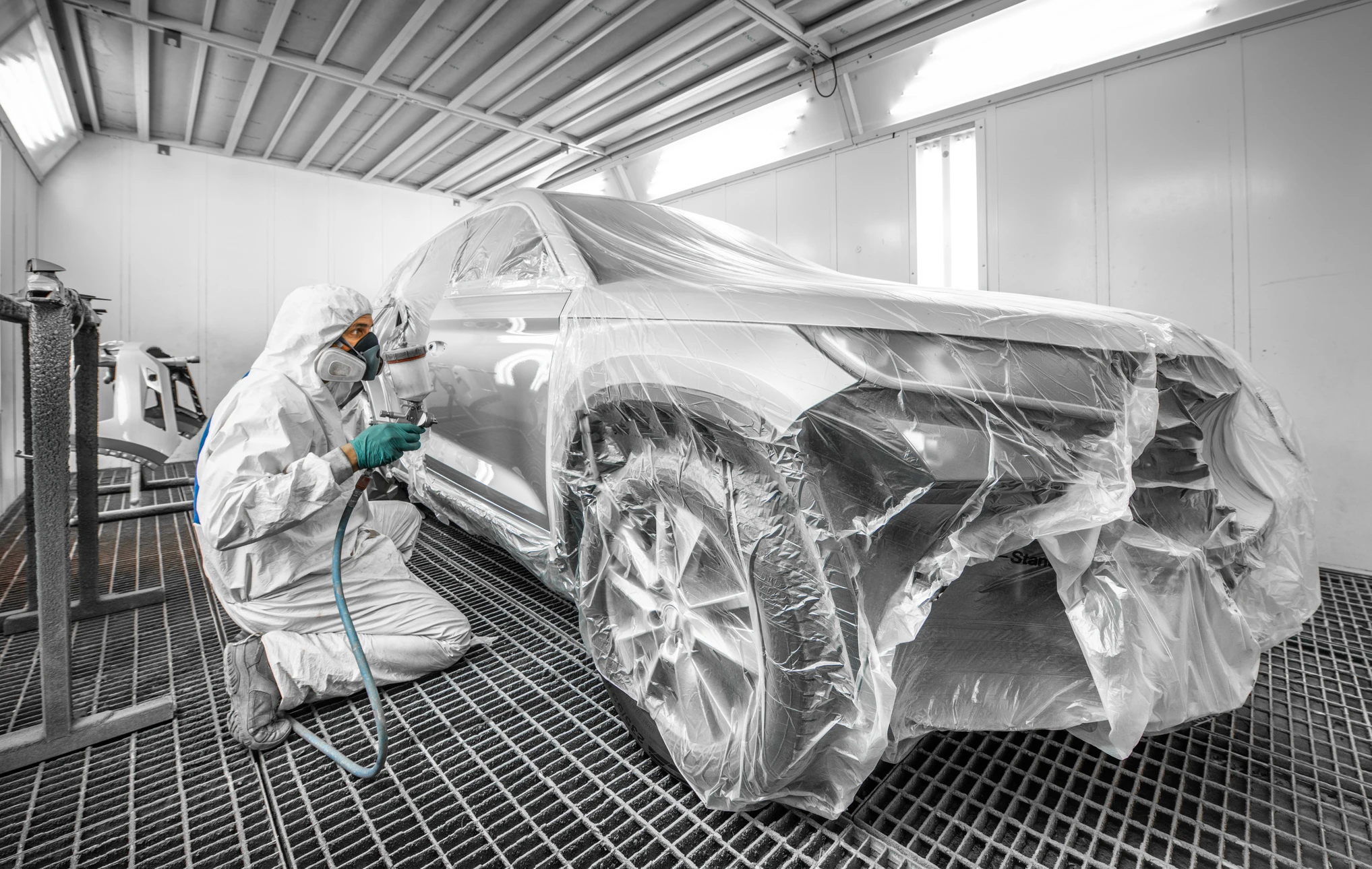 Atelier de carrosserie automobile avec technicien en combinaison de protection intégrale pulvérisant de la peinture sur véhicule entièrement bâché
