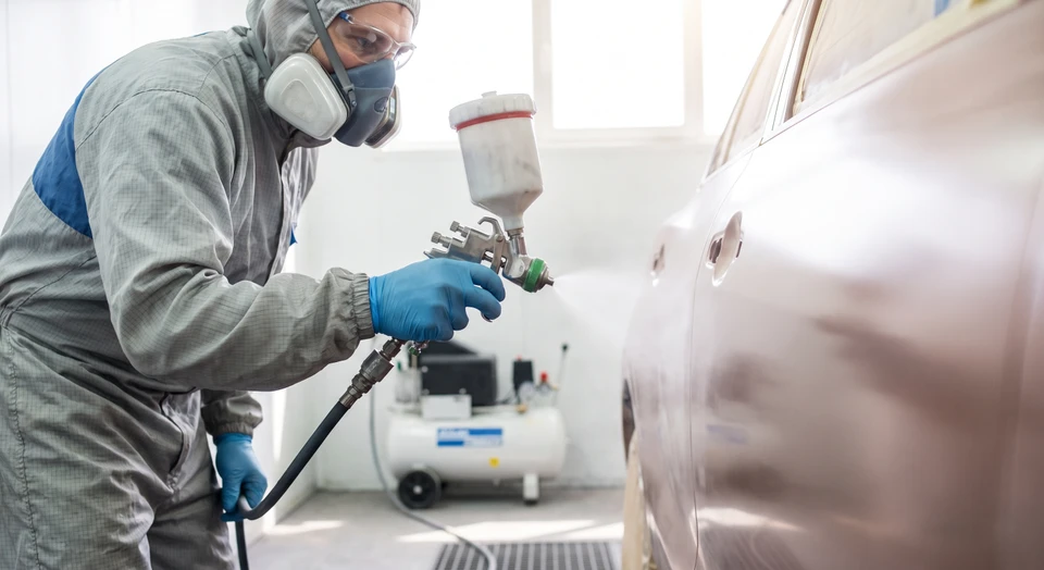 Gros plan d'un peintre carrossier dans un atelier automobile professionnel, utilisant un pistolet de pulvérisation pour appliquer de la peinture sur une carrosserie automobile. La scène montre précision et professionnalisme, avec l'équipement de peinture visible (pistolet HVLP, compresseur en arrière-plan). La teinte est en cours d'application sur une surface automobile lisse, illustrant la maîtrise technique et la colorimétrie. Lumière naturelle ou d'atelier professionnelle, équipement moderne et bien entretenu.